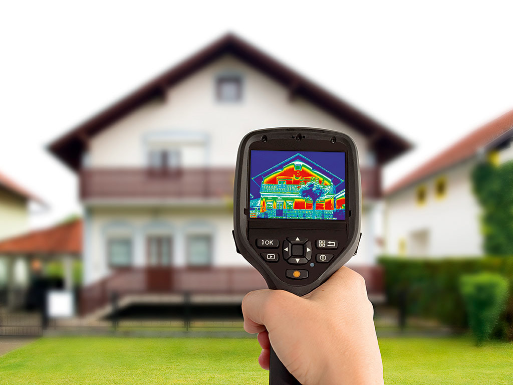 Picture of a hand holding a thermal camera with the camera showing a thermal image of the exterior of the home, with the visible view of the home in the background.